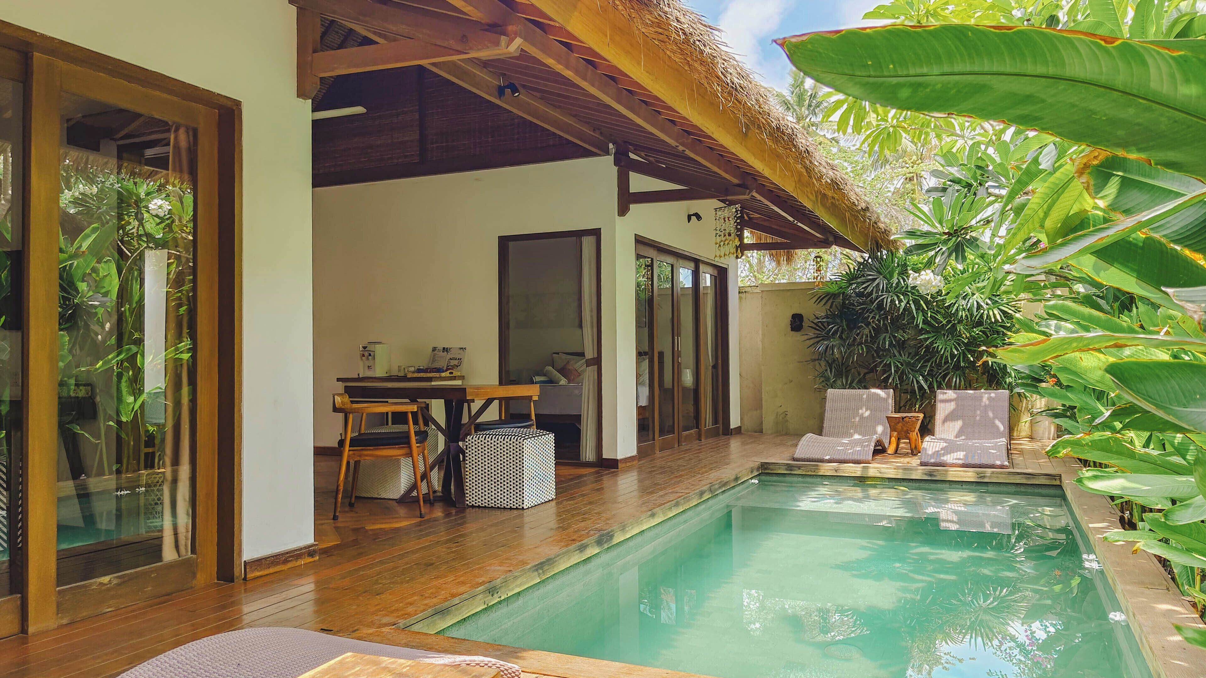 A pool area with wooden decking, a dining table with chairs, and a covered seating area with a thatched roof.