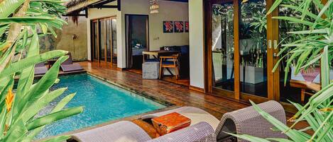 A poolside area with lounge chairs, a thatched roof structure, and tropical plants.