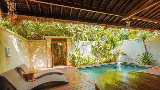 Zona junto a la piscina con sillones de descanso y una mesa de madera, rodeada de exuberante vegetación y un techo de paja.