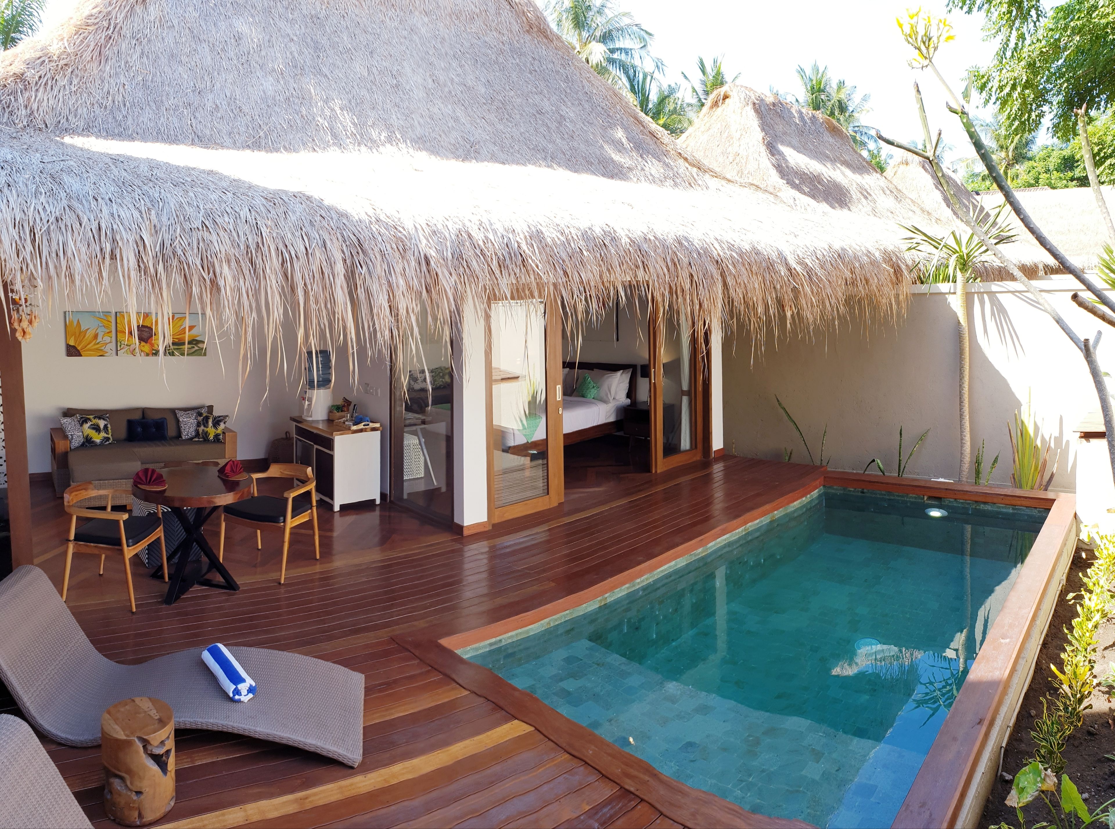 A thatched-roof bungalow with a pool, wooden deck, and a sofa with cushions.