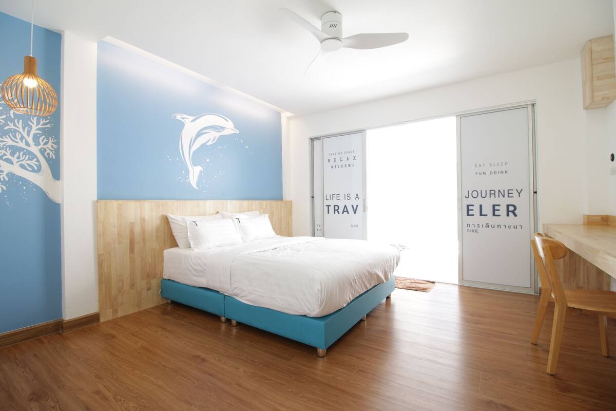 A modern bedroom with a bed, wooden flooring, a ceiling fan, and a wall with a dolphin mural.