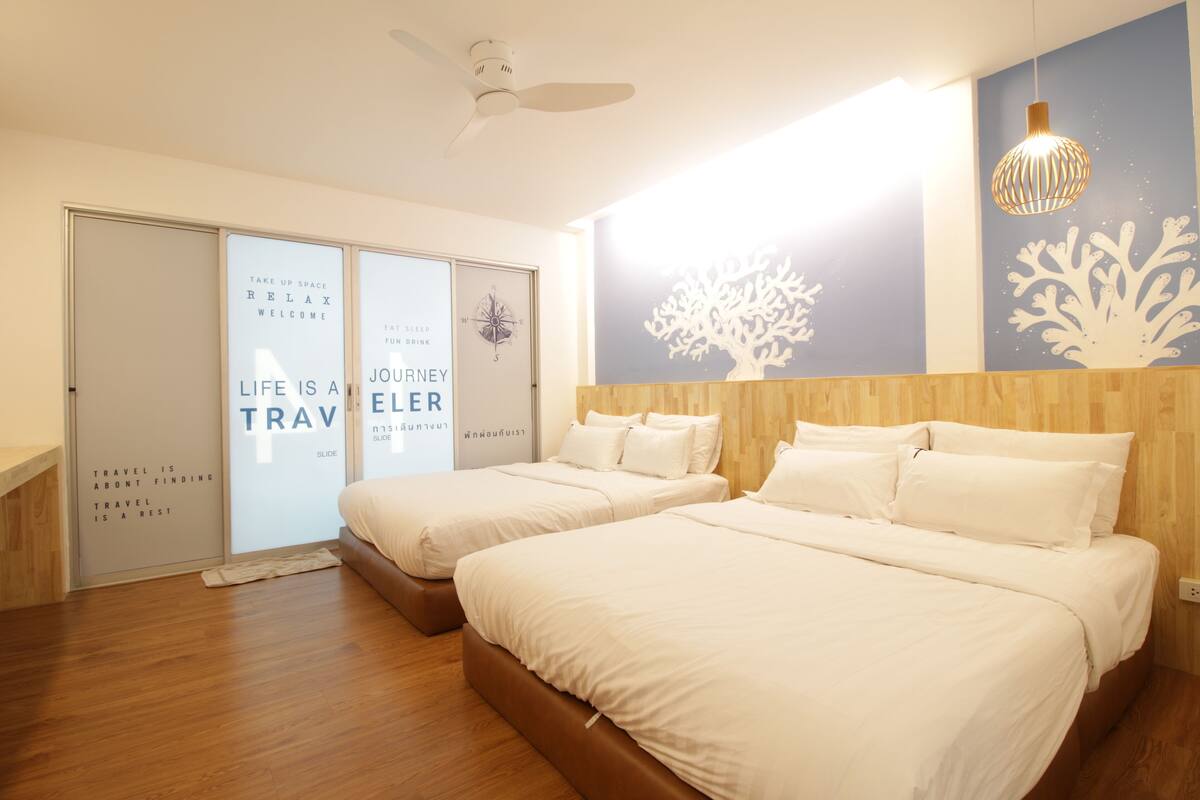 A hotel room with two beds, a ceiling fan, a wall with a tree design, and a light fixture.