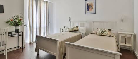 Una habitación con dos camas individuales, una mesita con una planta, una silla y un ventanal con cortinas.