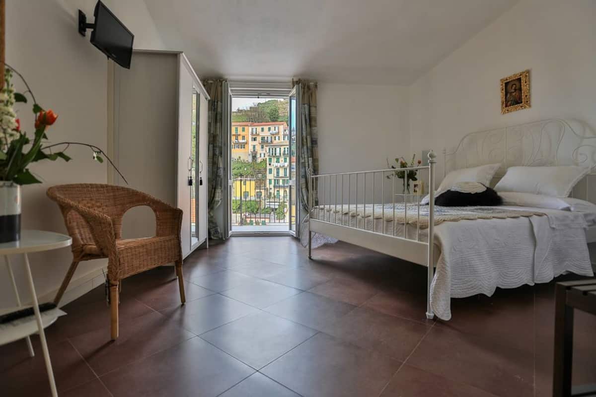 A bedroom with a white bed, a wicker chair, a small table, and a view of buildings through an open door.
