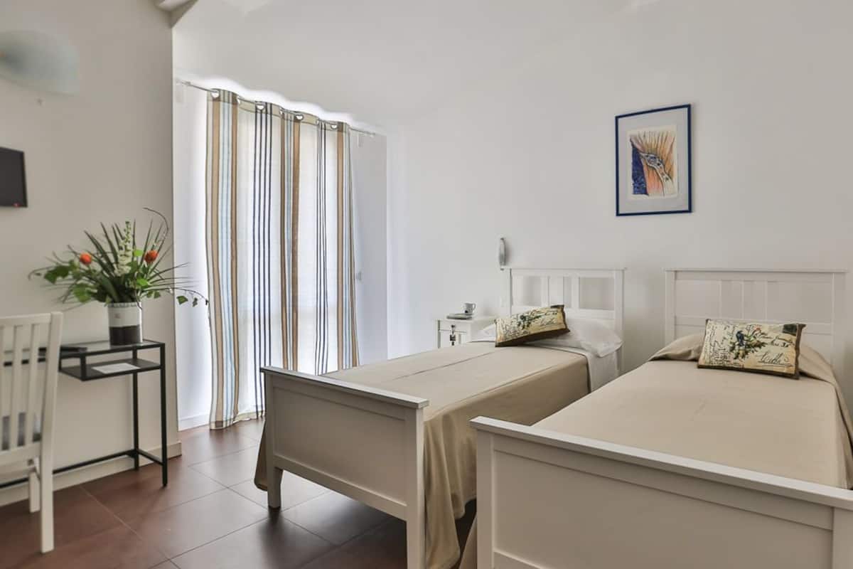 A room with two beds, a small table with a plant, a chair, a window with curtains, and a framed picture on the wall.