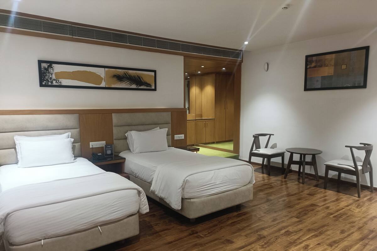 A hotel room with two beds, a small table, a chair, and a view of the corridor.
