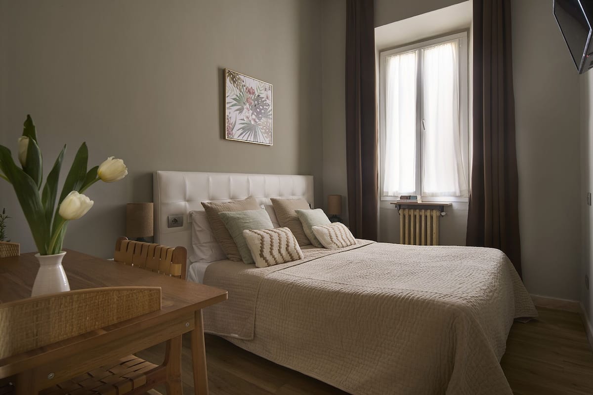 Une chambre à coucher avec un grand lit, une table de chevet, une lampe et une fenêtre avec des rideaux.