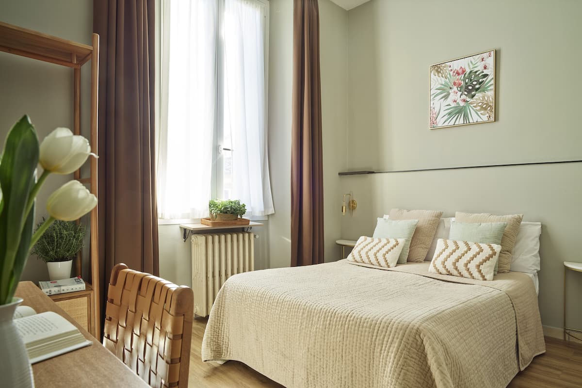 Une chambre à coucher comprenant un lit, une table de chevet, une chaise, une fenêtre avec des rideaux, un radiateur, une lampe fixée au mur et un tableau encadré.