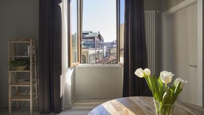 A room with a large window, a wooden table with a vase of white flowers, a wooden ladder shelf with books and a plant, and a view of buildings outside. - B&B My Way (Florence)