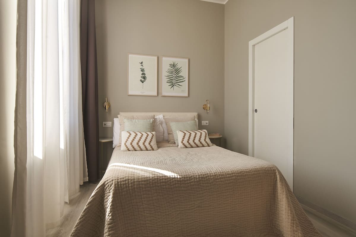Une chambre à coucher avec un lit, deux reproductions botaniques encadrées accrochées au mur, une porte et une fenêtre avec des rideaux.
