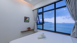 A hotel room with a large window offering a view of the sea, a neatly made bed, a small shelf with flowers, and a framed picture on the wall. - Handy Beachfront Apartment (Bac Nha Trang)