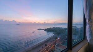 A coastal view from a window overlooking a beach with calm waters, a road, and buildings in the distance. - Handy Beachfront Apartment (Bac Nha Trang)