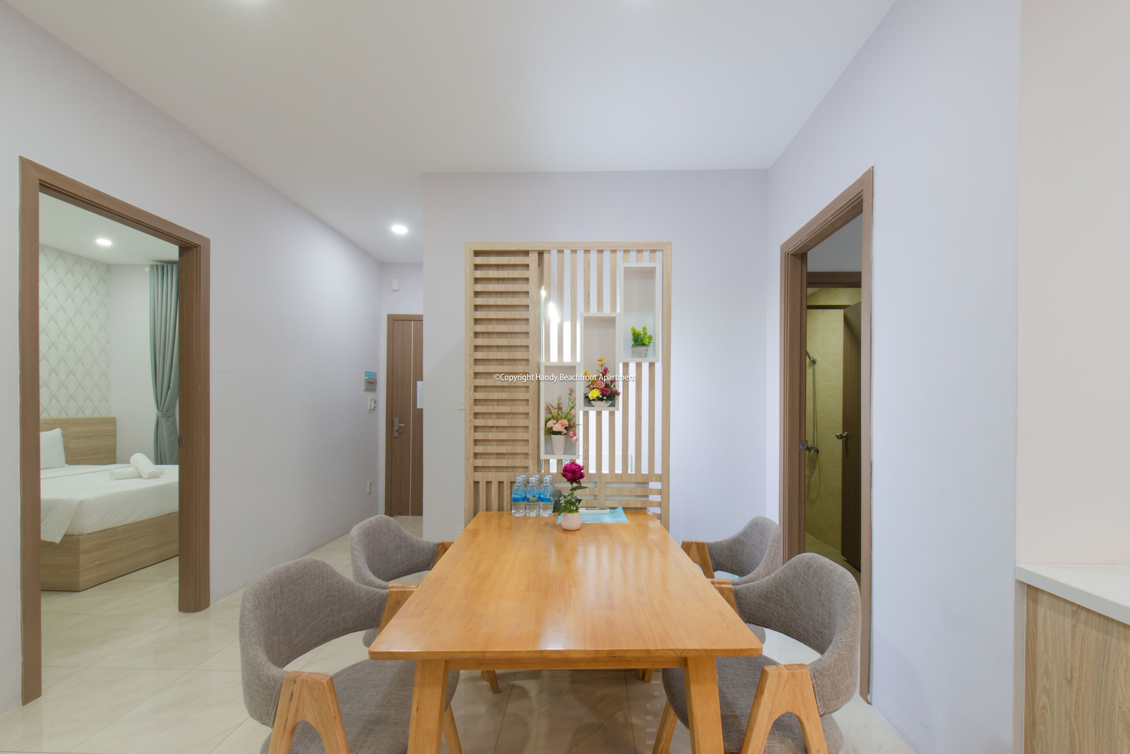A modern hotel room with a wooden dining table, grey upholstered chairs, a small potted plant, and a decorative wall panel.