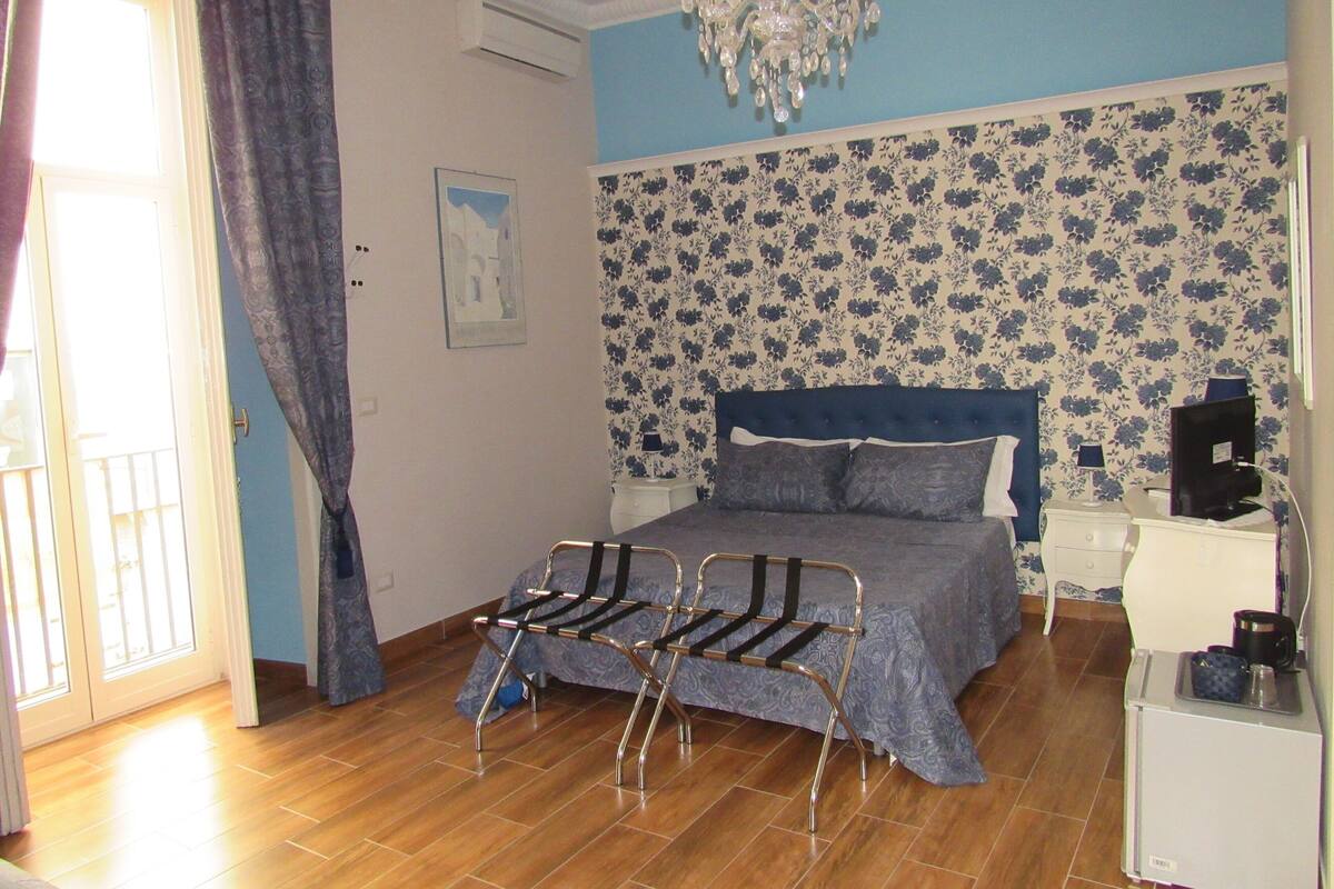 A bedroom with a bed, a nightstand, a dresser, a chair, a chandelier, and a window with curtains.
