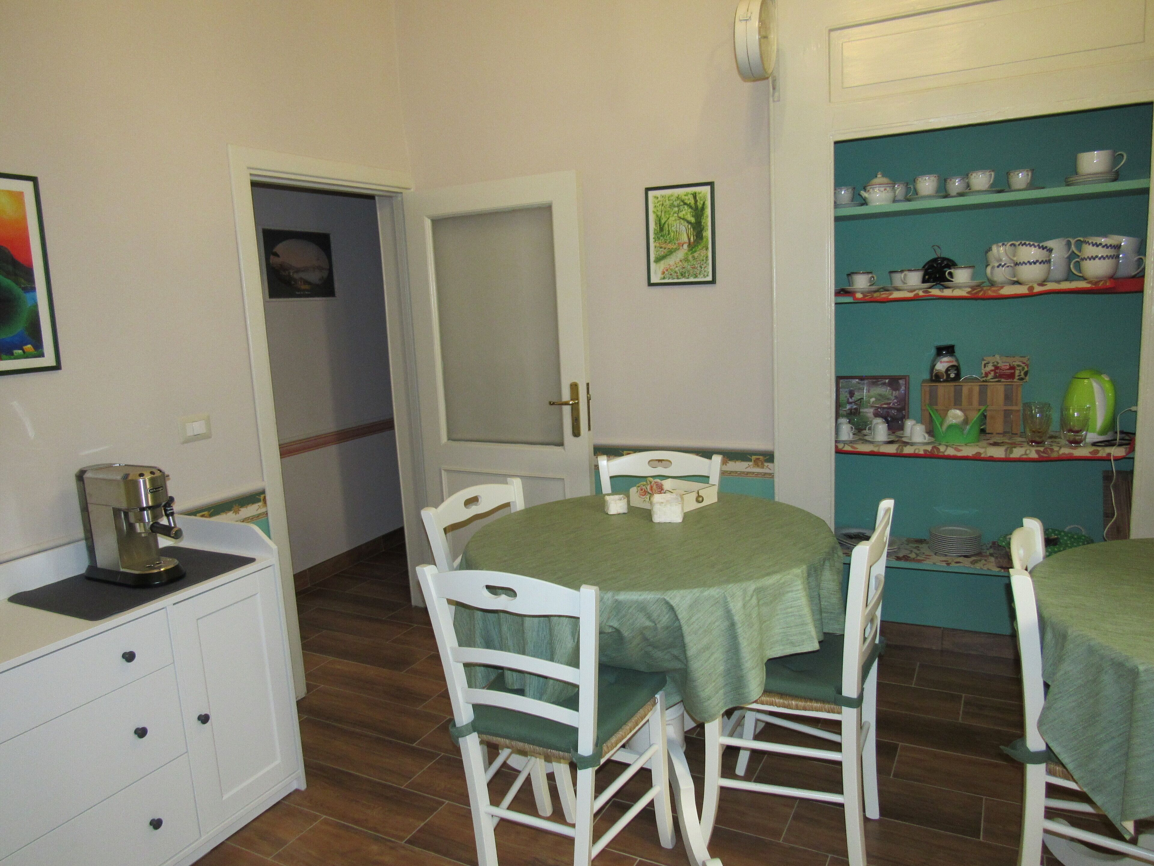 A dining area with a round table covered by a green tablecloth, surrounded by white chairs. There is a white cabinet with drawers to the left.
