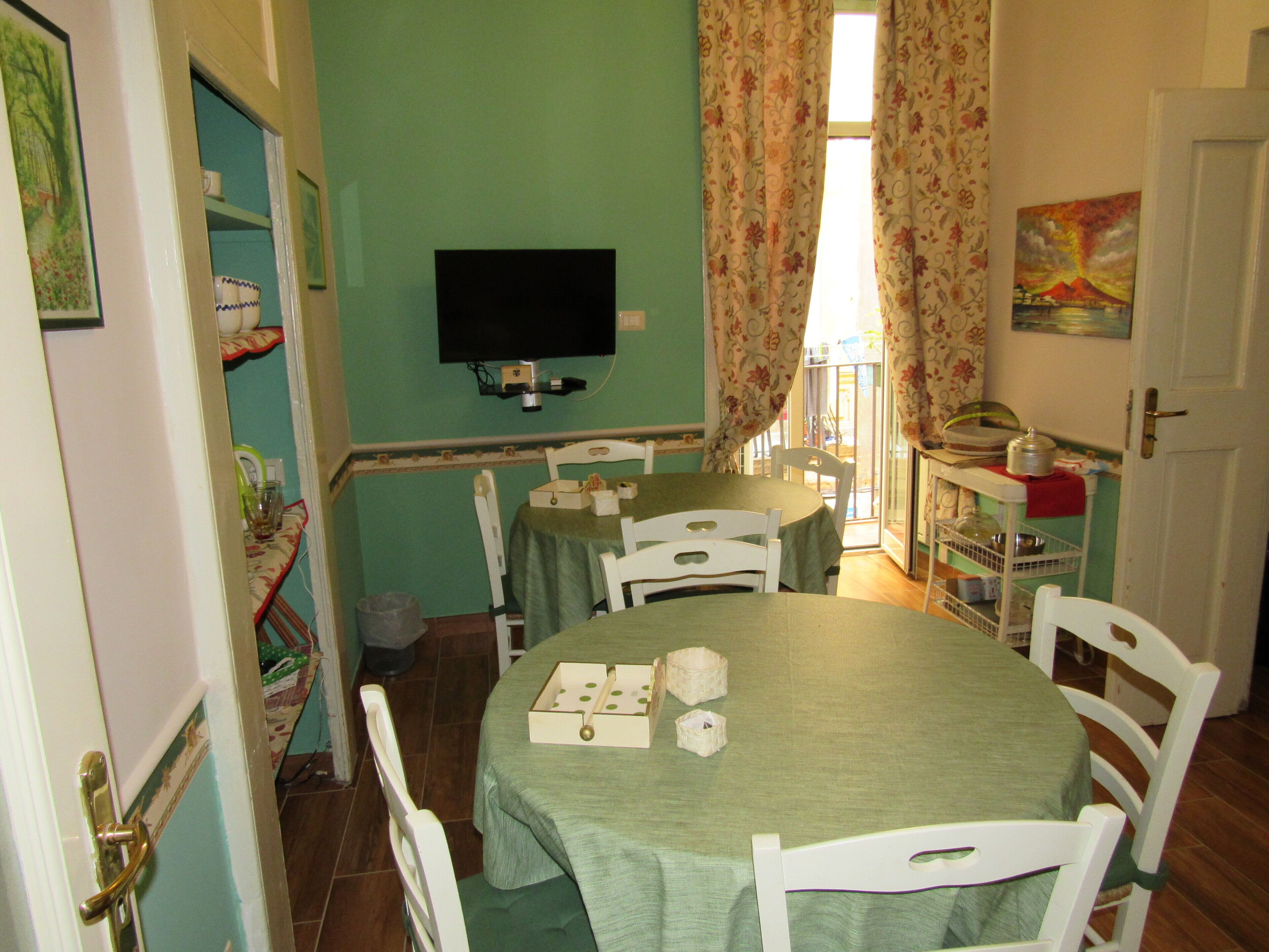 Un'area pranzo con un tavolo verde, sedie bianche, una televisione a parete e un quadro appeso al muro.