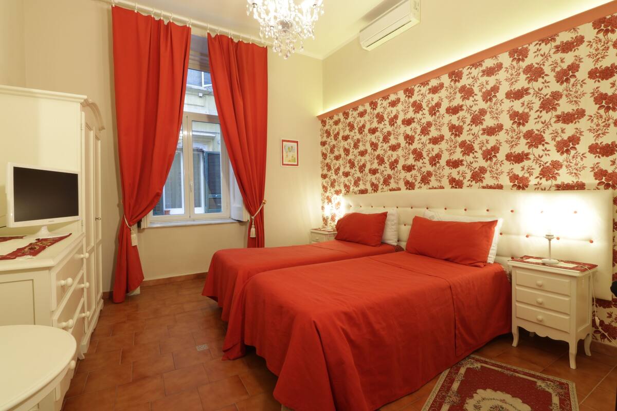 A bedroom with two beds, a TV, a dresser, and a window with red curtains.
