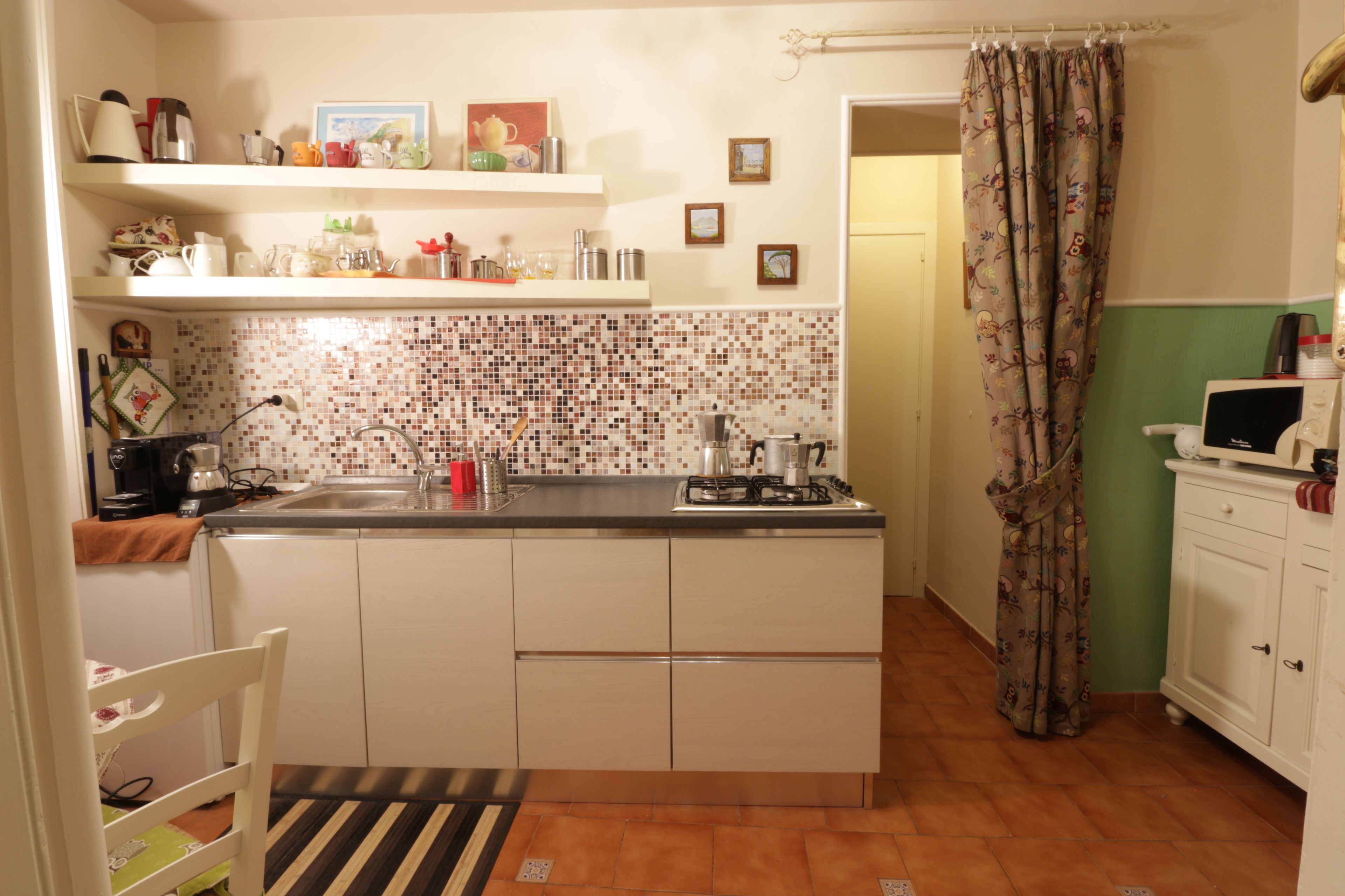 Una cucina compatta con rivestimento in piastrelle, fornello a gas, lavello e forno a microonde. Sopra il bancone, degli scaffali contengono diversi oggetti da cucina.