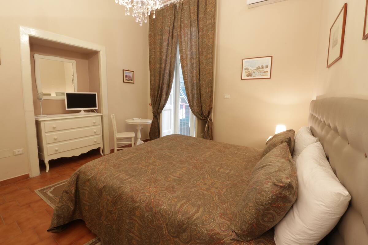 A bedroom with a bed, a dresser, a mirror, a television, and a window with curtains.