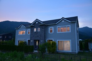 A two-story house with a stone facade and large windows, surrounded by a fenced yard and mountains in the background. - Kawaguchiko Cottage minami (Fujikawaguchiko)