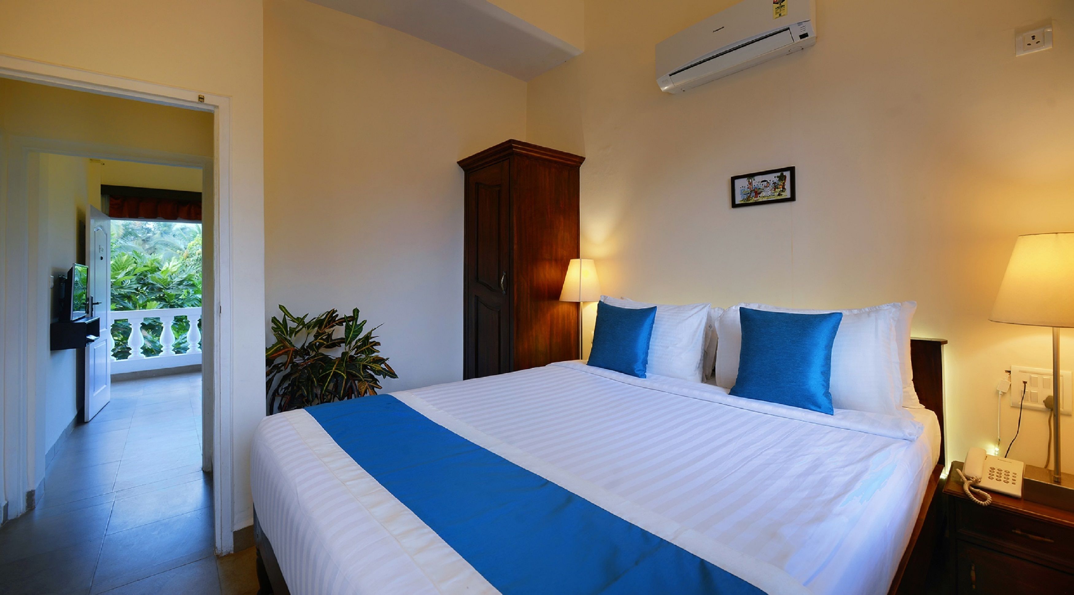 Quarto de hotel com cama, mesa de cabeceira, luminária, ar condicionado e vista para uma varanda com plantas.