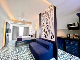 Habitación de hotel con cama, sofá, separador decorativo, microondas y pequeña cocina.