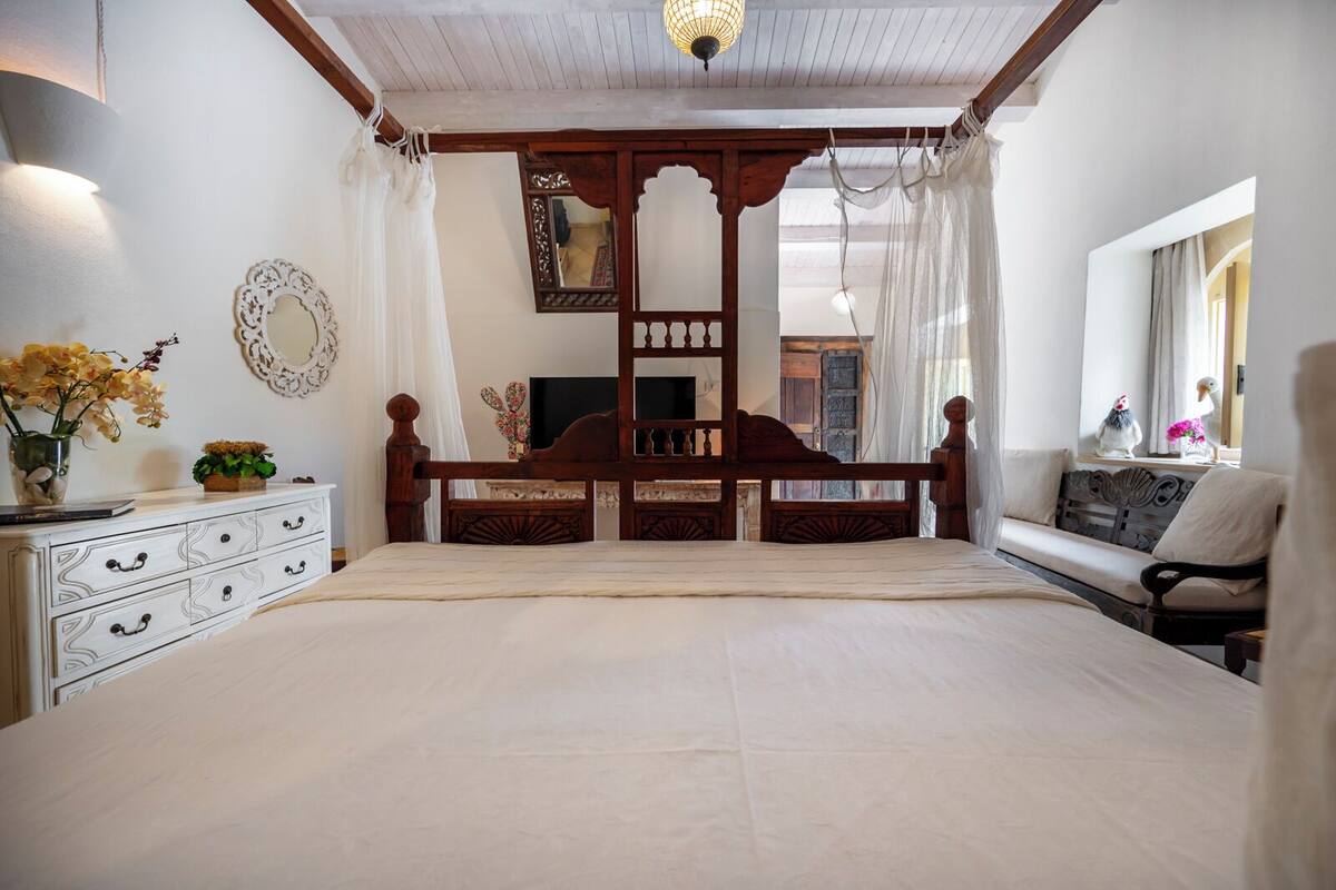 A spacious bedroom with a large bed, wooden headboard, a bedside table with a vase of flowers, and a window with curtains.
