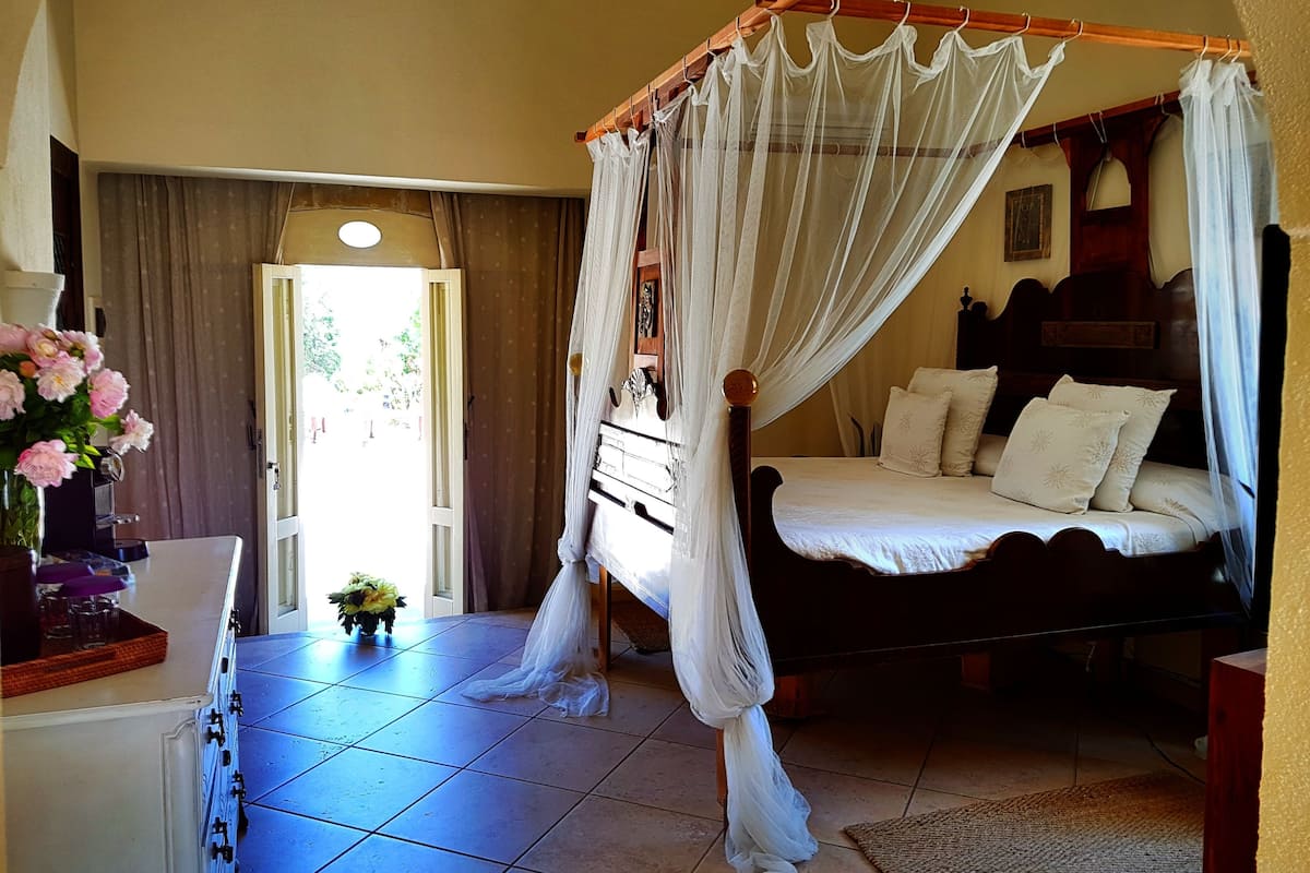 A bedroom with a four-poster bed, a bedside table, a dresser, and a window with curtains.