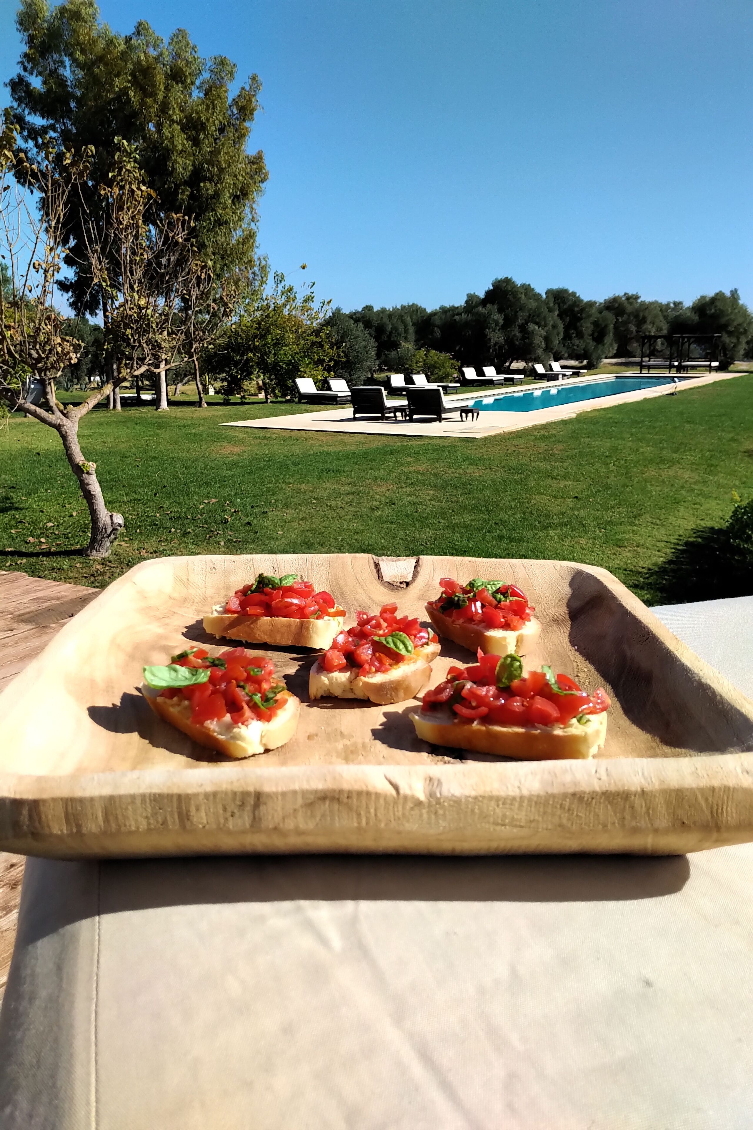 A wooden tray with food items on a table, overlooking a pool and a grassy area with trees and lounge chairs.