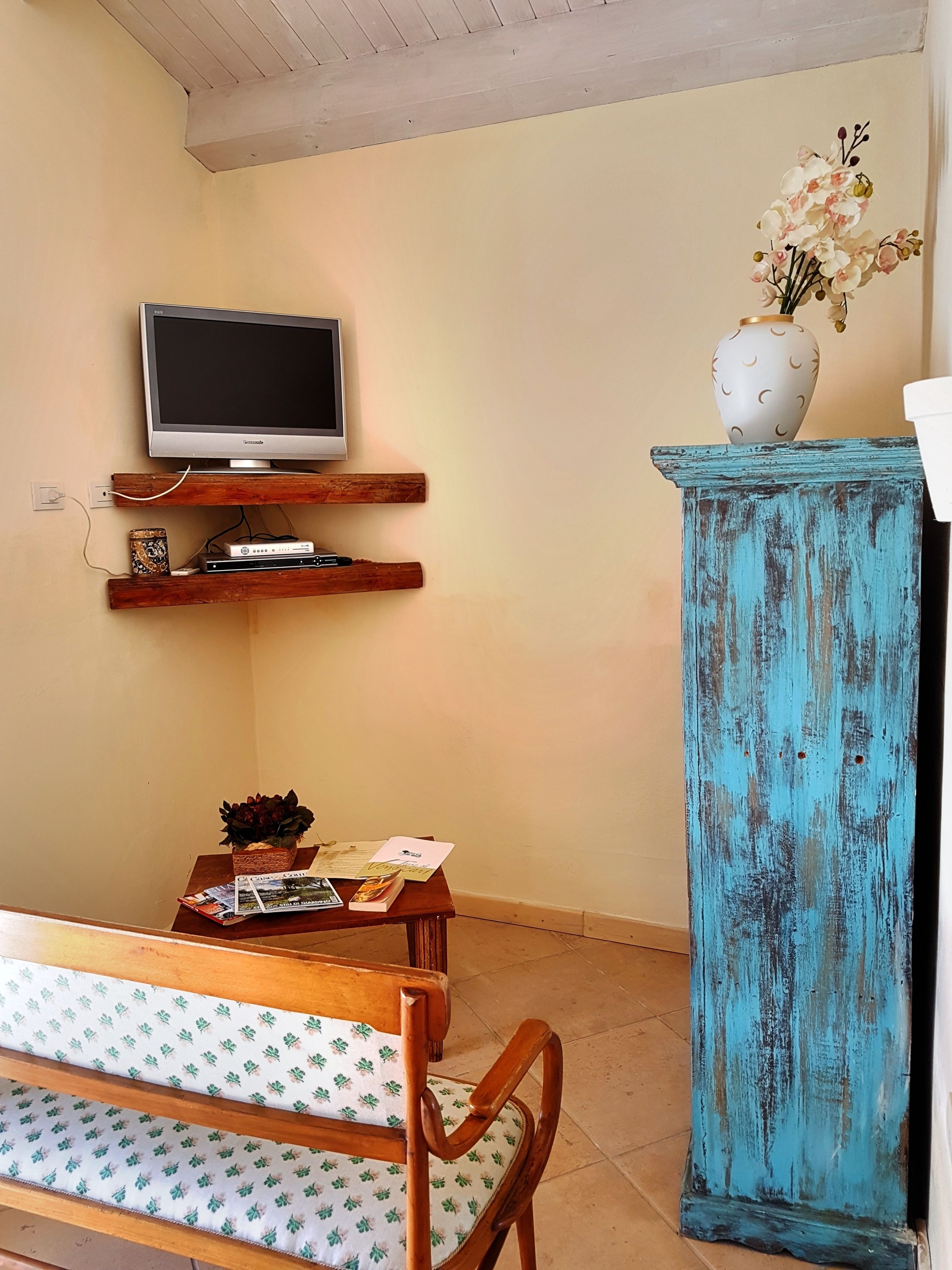 Una sala de estar con un televisor en un estante de madera, un armario azul, un banco de madera y una mesita con revistas y una planta.