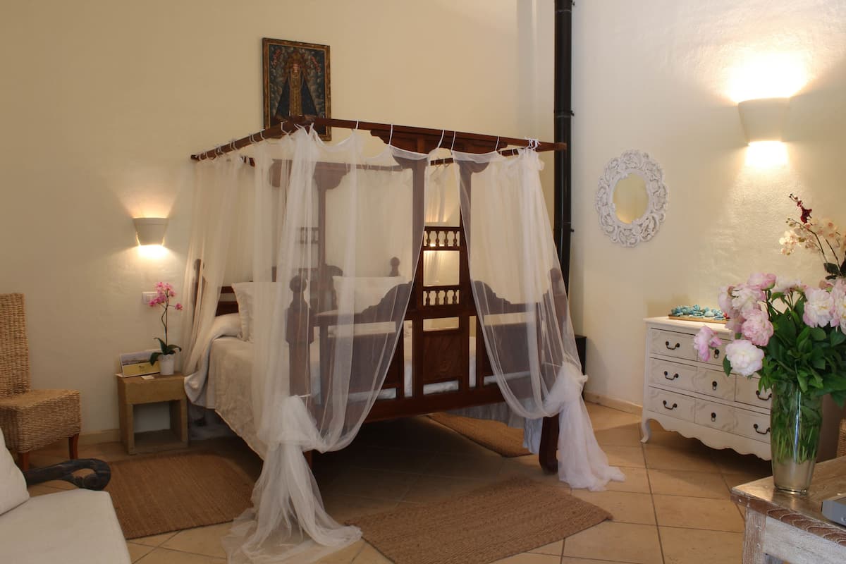 A bedroom with a four-poster bed, a bedside table with a lamp, a dresser, and a vase with flowers.