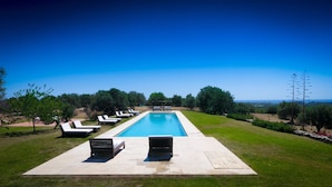 Une piscine rectangulaire à l’eau d’un bleu limpide, entourée d’une terrasse en béton. Des chaises longues sont alignées de part et d’autre de la piscine, et quelques bancs sont placés au centre. La terrasse est bordée d’un petit muret en pierre, et en arrière-plan, on aperçoit une zone herbeuse avec des arbres et un ciel dégagé.