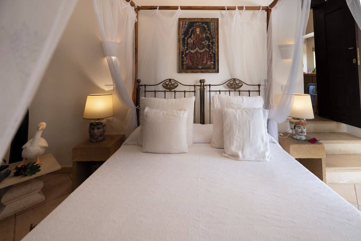 A four-poster bed with white linens, two bedside lamps, and a painting above the headboard.