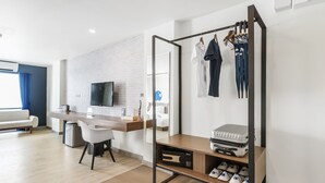 Une chambre d’hôtel moderne dotée d’un bureau en bois, d’un téléviseur à écran plat, d’un coin salon avec un canapé blanc et d’un portant à vêtements.
