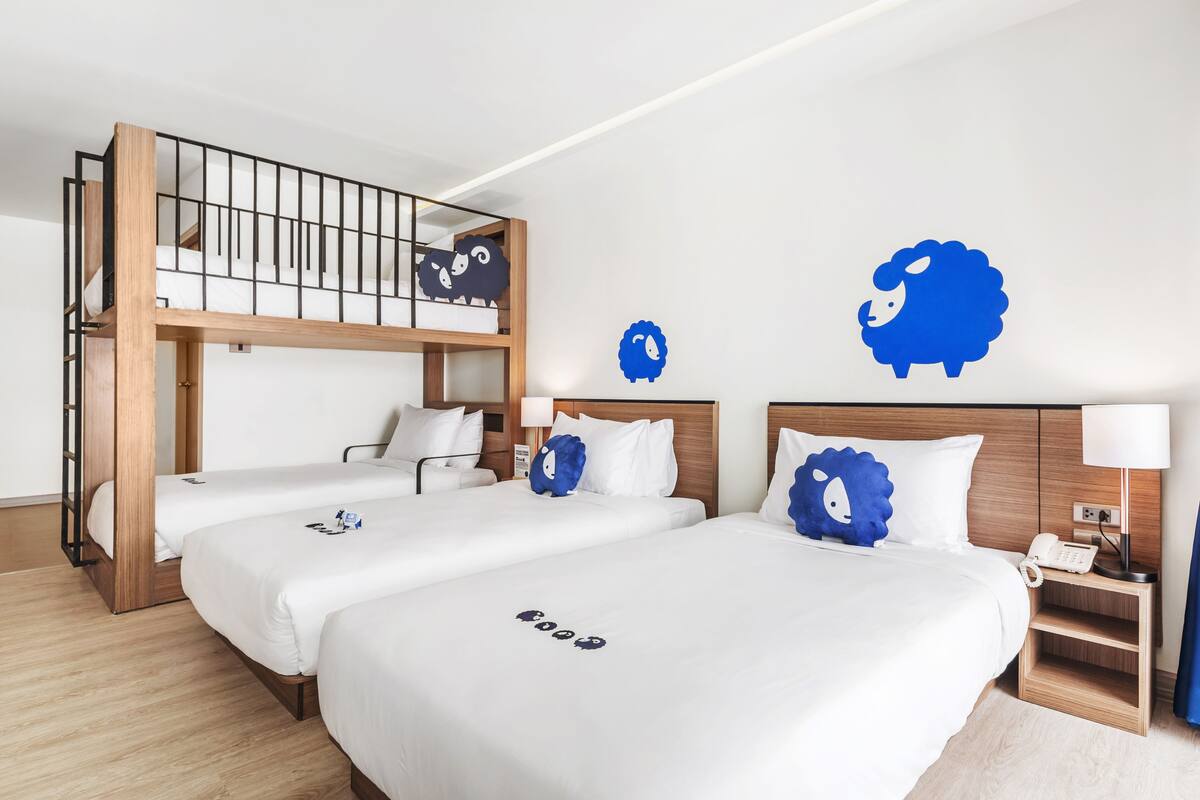 Ein Hotelzimmer mit Etagenbett, zwei Einzelbetten, einem Holzkopfteil, einem Nachttisch mit Lampe und dekorativer Wandkunst mit blauen Schafen.