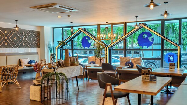 Un café moderne avec des tables et des chaises en bois, un canapé et une grande fenêtre donnant sur un espace verdoyant et des voitures garées.