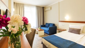 A hotel room with a bed, a blue sofa, a wooden nightstand, a desk with a vase of flowers, and a window with curtains. - Ljuljak (Golden Sands)