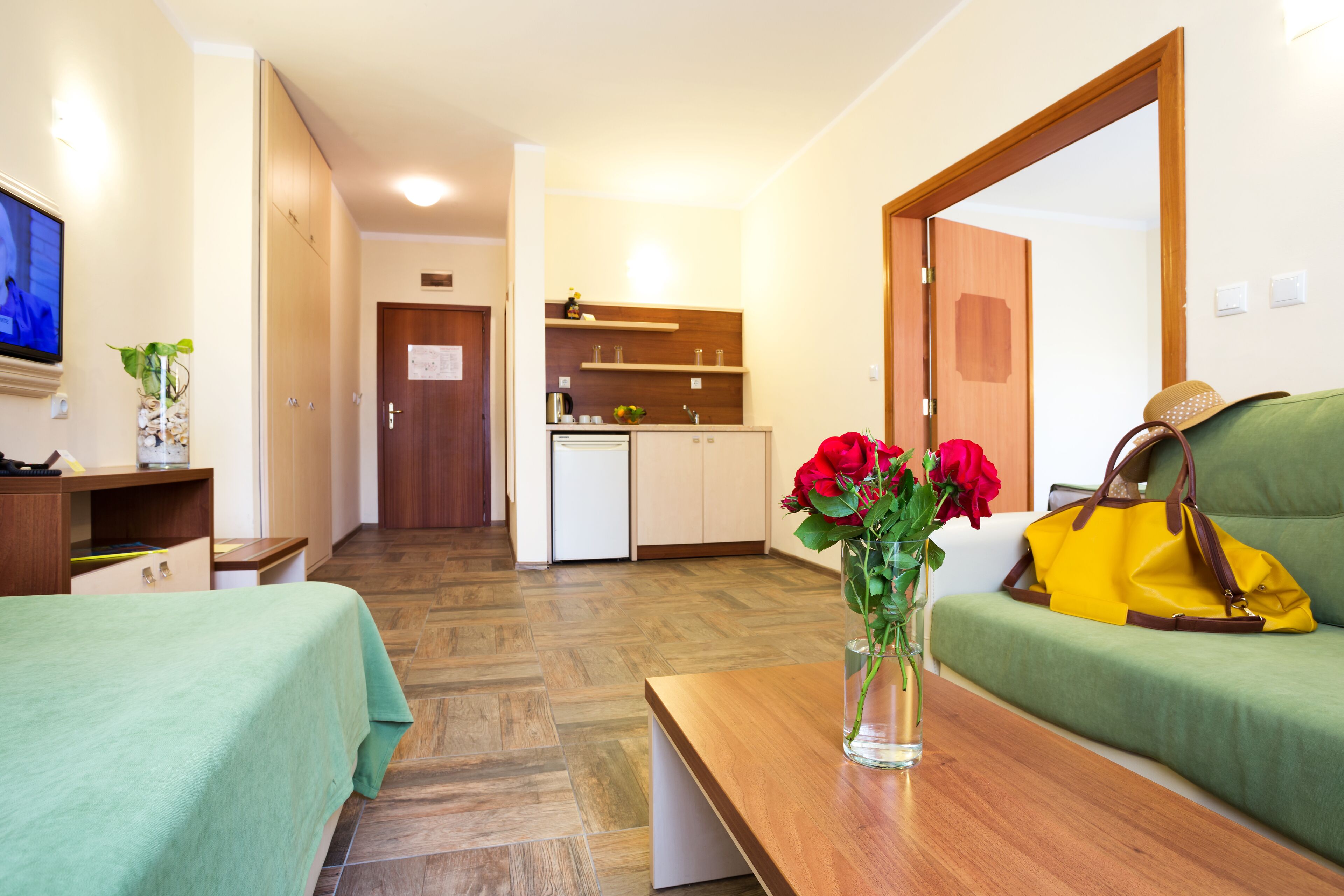 Habitación de hotel con una cama, una mesa de madera con un florero, un sofá, un televisor y una kitchenette al fondo.