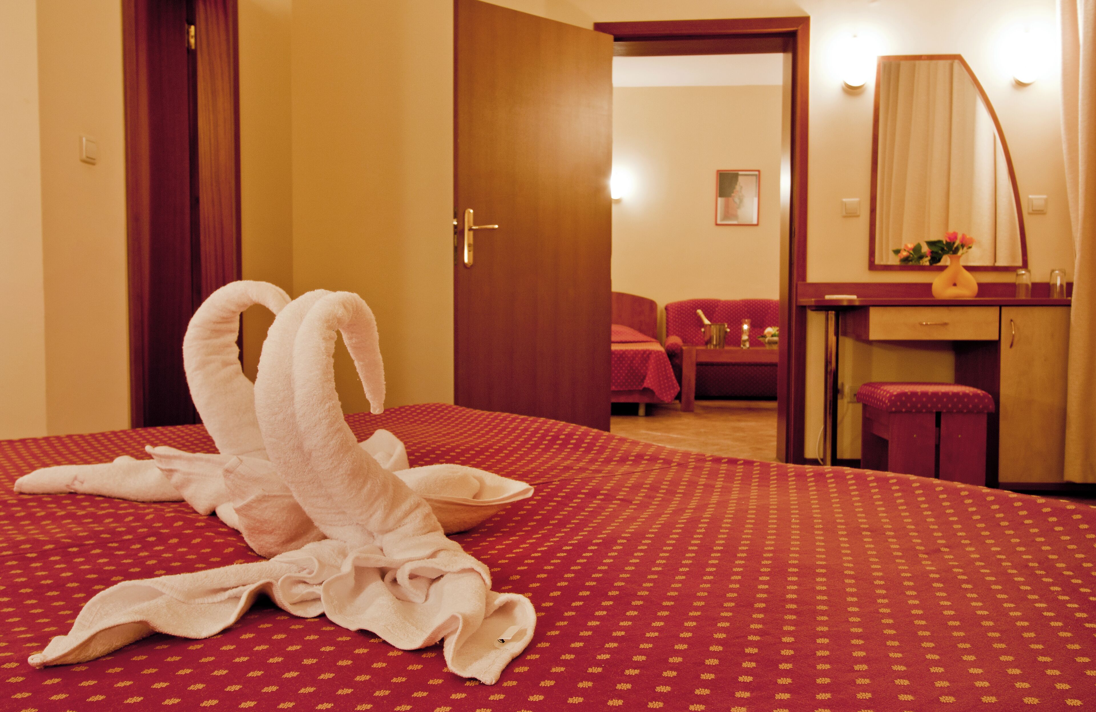 Ein Hotelzimmer mit rotem, gemustertem Bettbezug, einer Holztür, einem roten Sessel, einem kleinen Tisch mit einer Vase und einem Spiegel an der Wand.