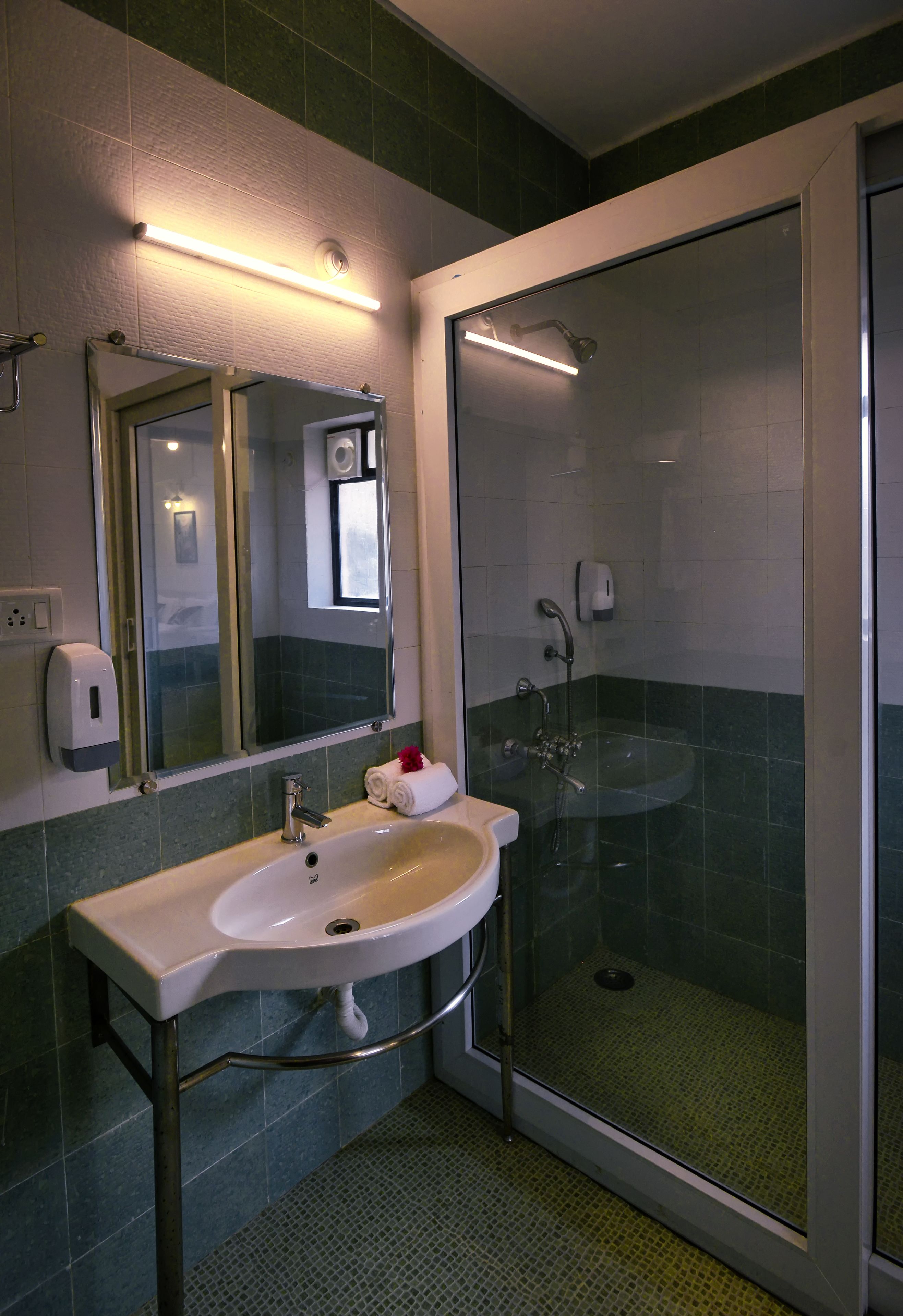 A bathroom with a glass-enclosed shower, a sink with a mirror above it, and a soap dispenser on the wall.