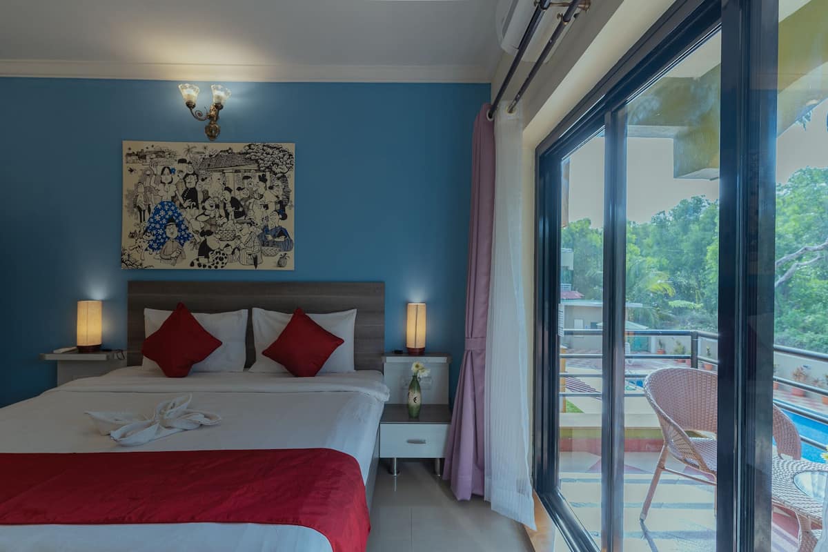 Habitación de hotel con una cama grande, dos lámparas de noche, una mesita de noche, una manta roja, un jarrón con flores y un ventanal con vistas a un balcón y árboles.