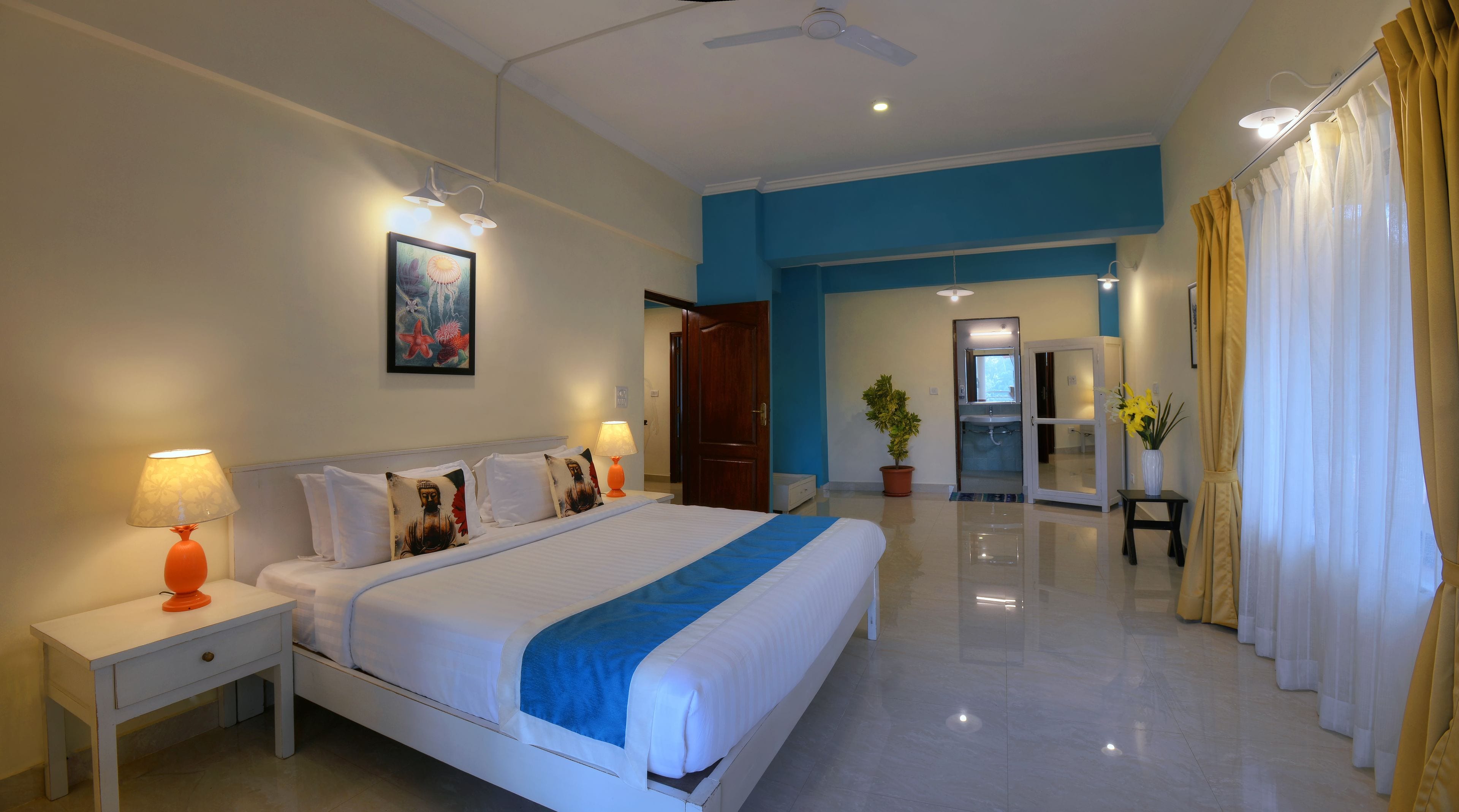 Habitación de hotel con una cama grande, lámparas de noche, ventilador de techo, baño visible a través de una puerta abierta y una planta en maceta.