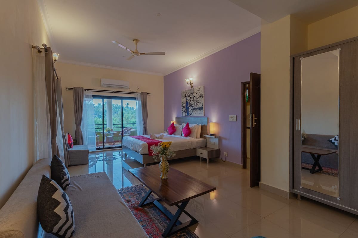 Habitación de hotel con cama, sofá, mesa de comedor y balcón con vista a zonas verdes.