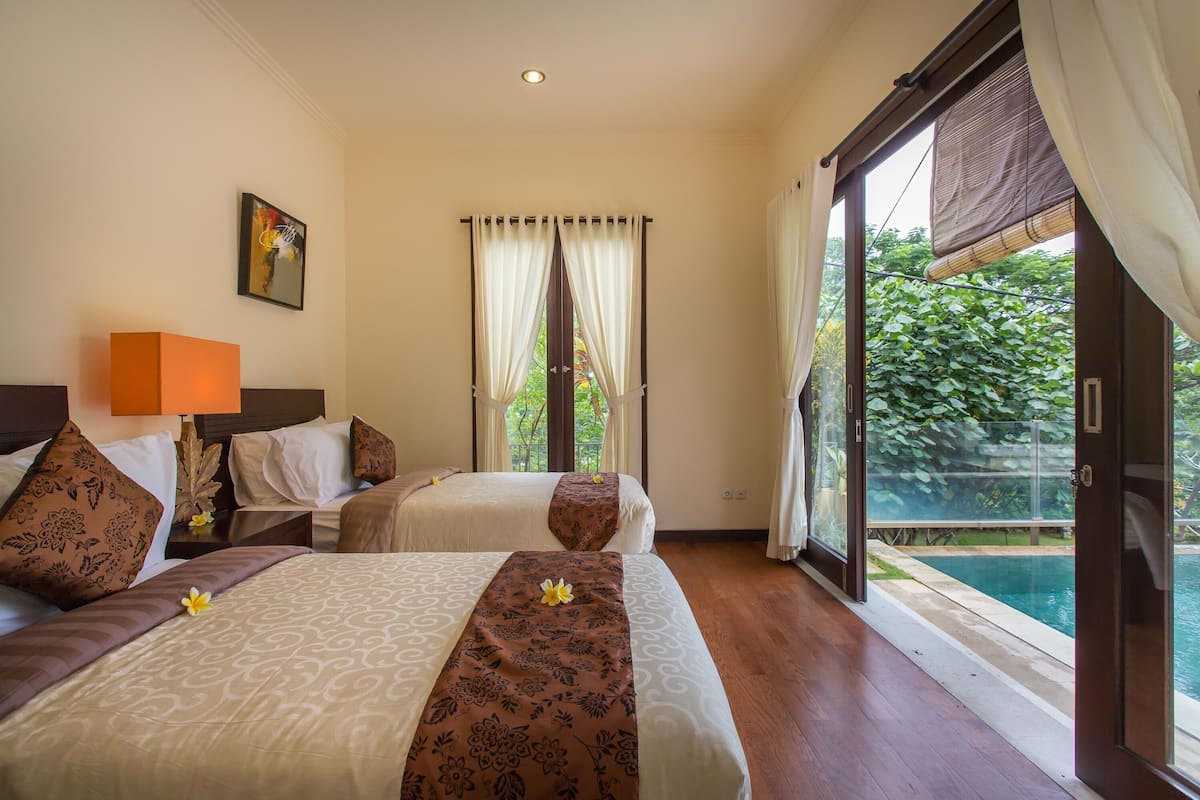 A hotel room with two beds, a large window with curtains, a view of greenery, and a pool.