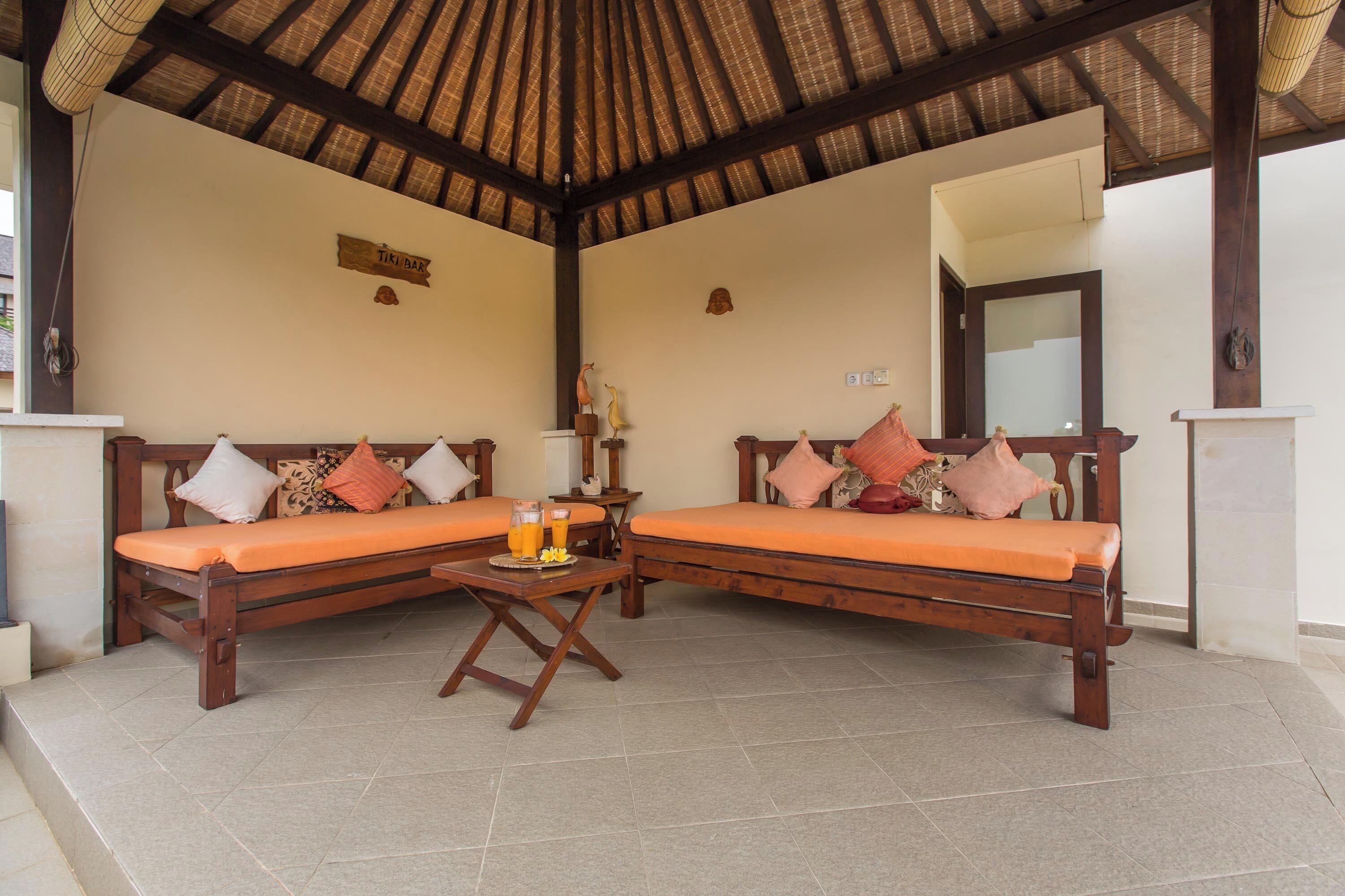 A room with two wooden sofas with orange cushions, a small table with a tray of orange drinks, and a thatched roof ceiling.
