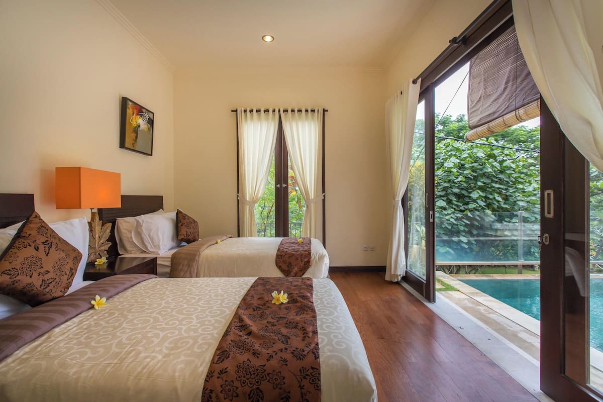 A hotel room with two beds, a large window with curtains, a painting on the wall, and a view of a swimming pool.