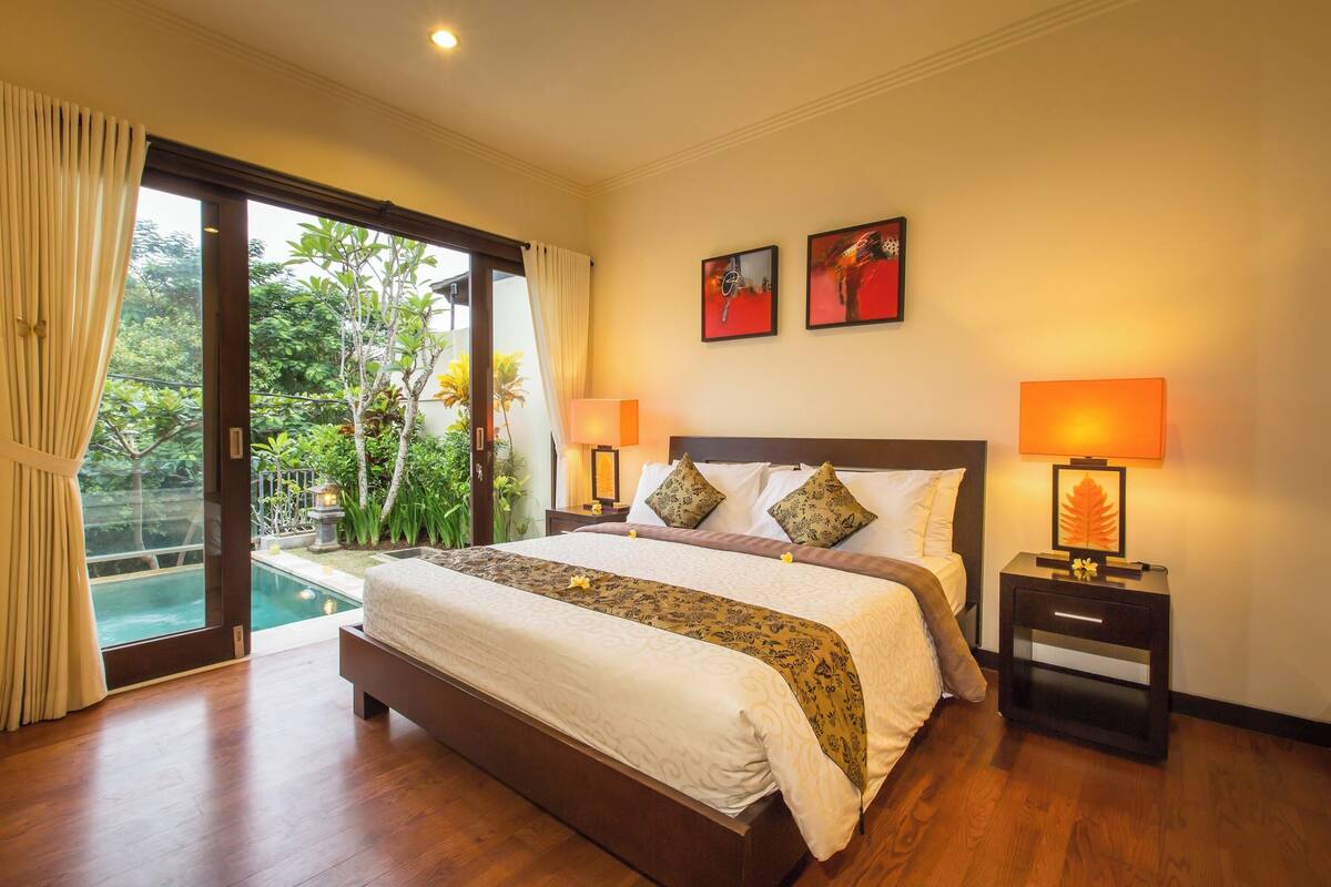 A spacious bedroom with a large bed, two bedside tables with lamps, a view of a swimming pool, and two framed artworks on the wall.