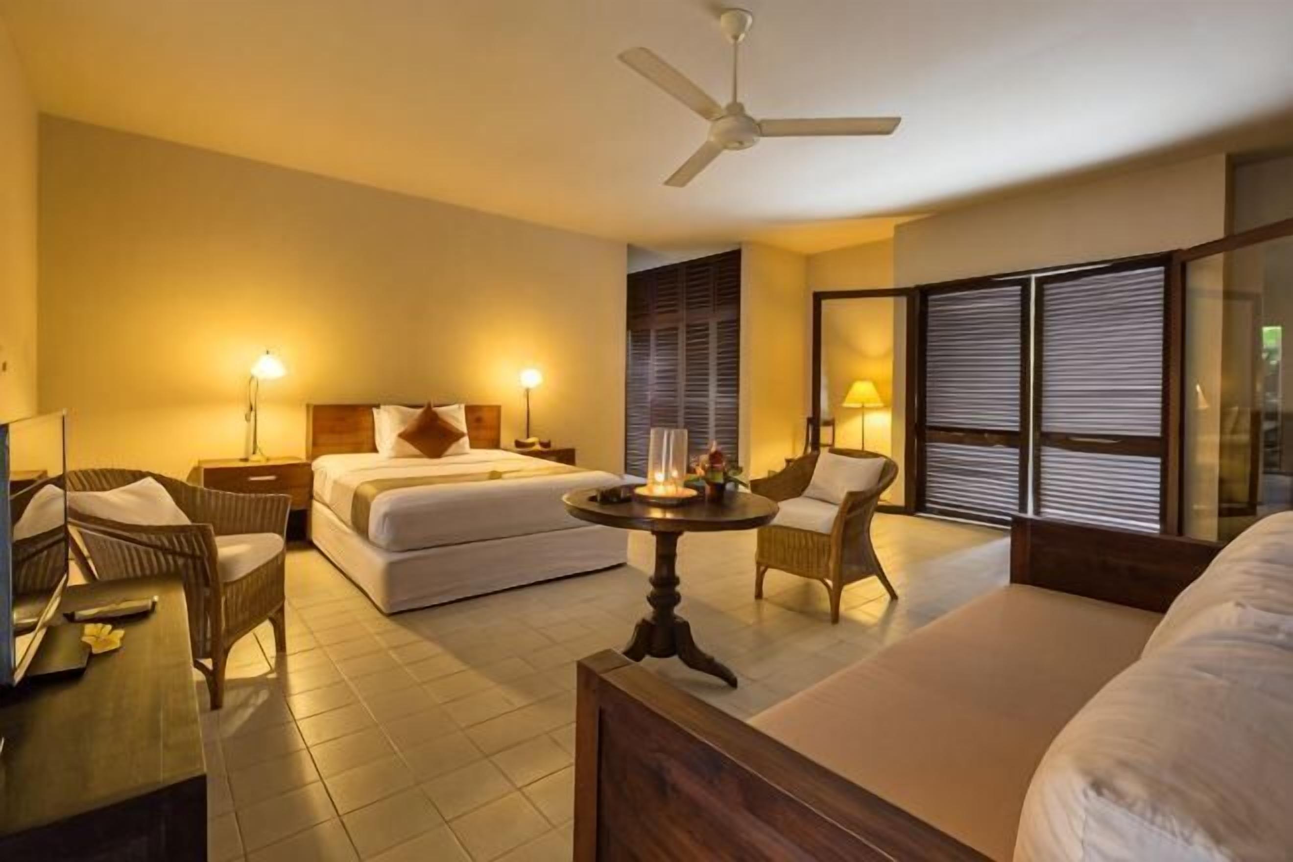 Quarto de hotel com cama, duas cadeiras, uma mesa, ventilador de teto e uma janela grande com persianas.
