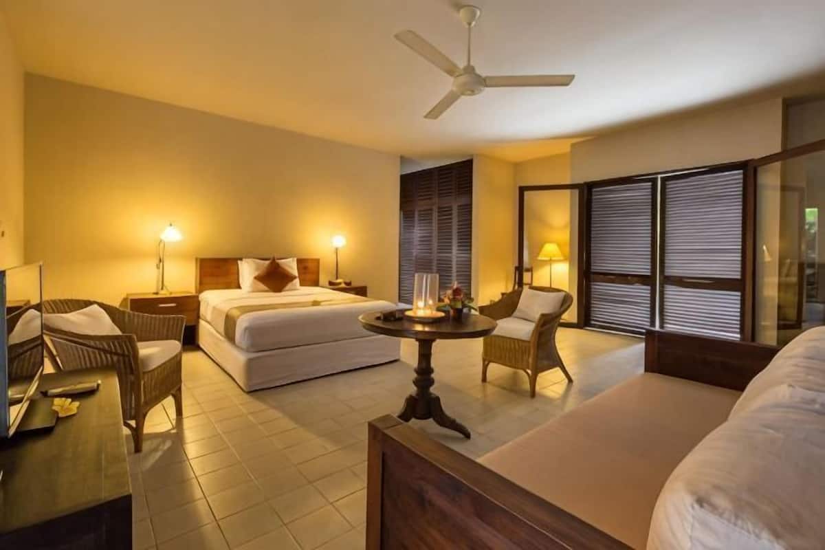 A hotel room with a bed, two chairs, a table, a ceiling fan, and a large window with blinds.