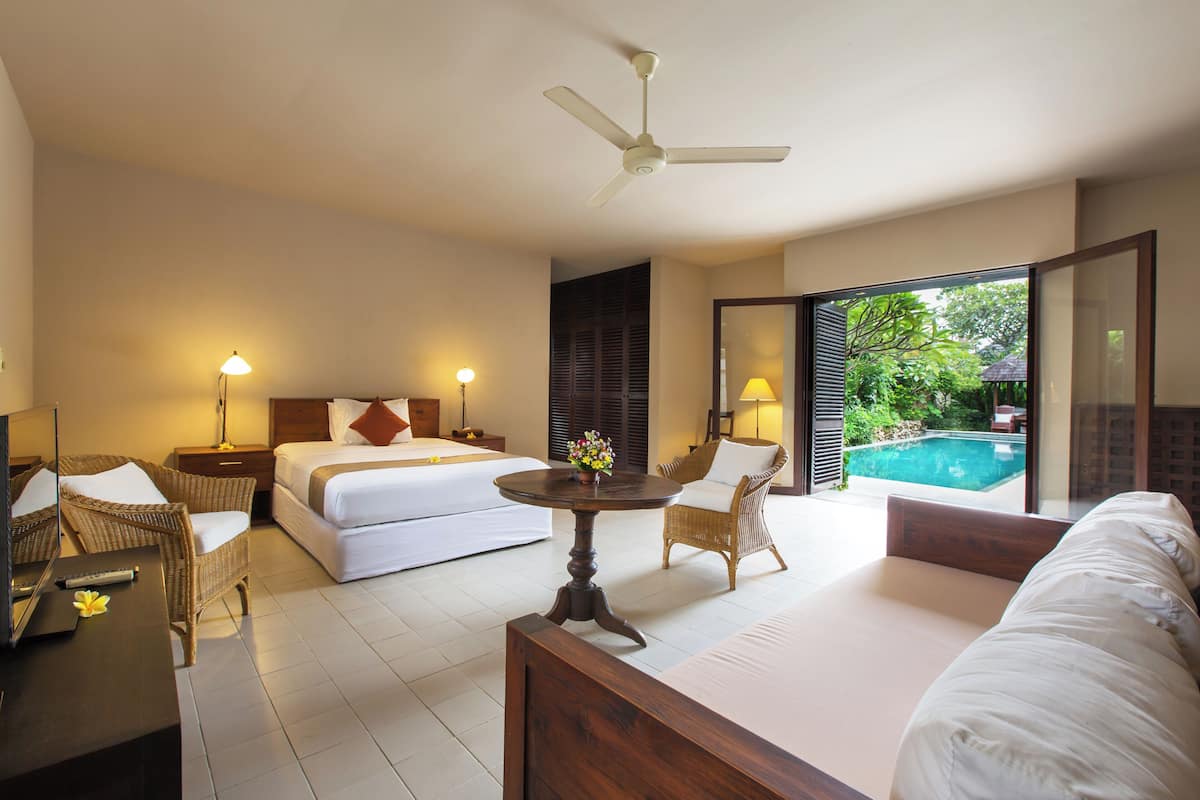 A hotel room with two beds, a dining table, wicker chairs, a ceiling fan, and a view of a swimming pool.